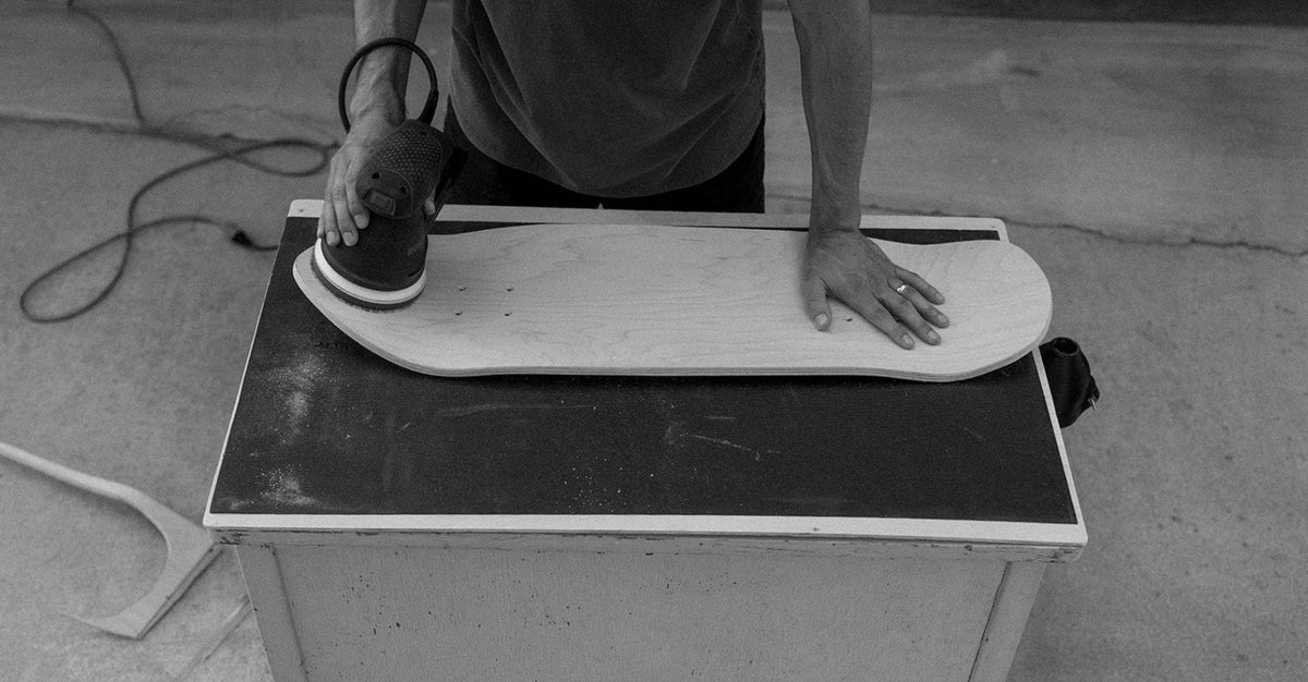 A black and white photo shows a person using a circular saw to cut a skateboard deck resting on a workbench in a workshop setting.