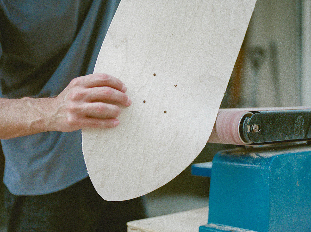 A person using a belt sander to shape a light-colored wooden skateboard deck. The person is wearing a blue-gray t-shirt and dark pants, and the sander is mounted on a workbench.