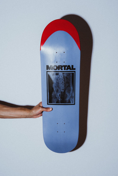 A light blue skateboard deck with a red top strip and a black-and-white X-ray graphic, held by a person's hand against a pale gray wall.